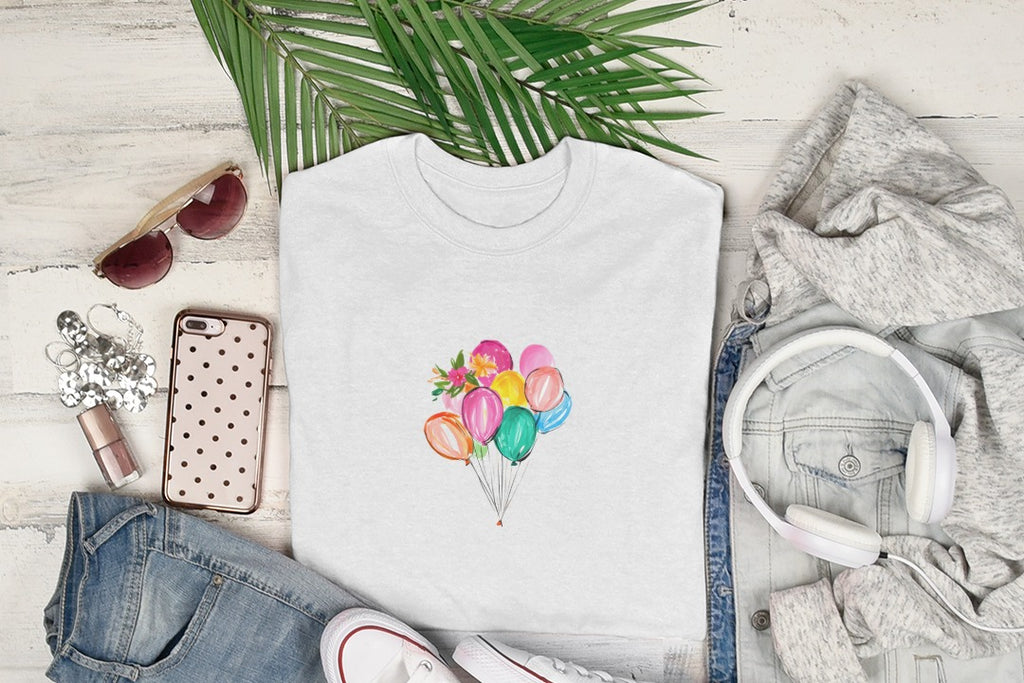 White t-shirt with colorful watercolor balloons graphic, laid flat with sunglasses, phone, jeans, and headphones.