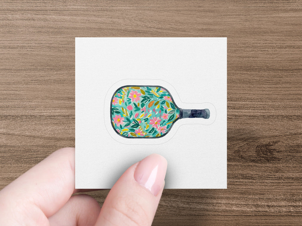 Pickleball paddle with floral pattern held by a hand.
