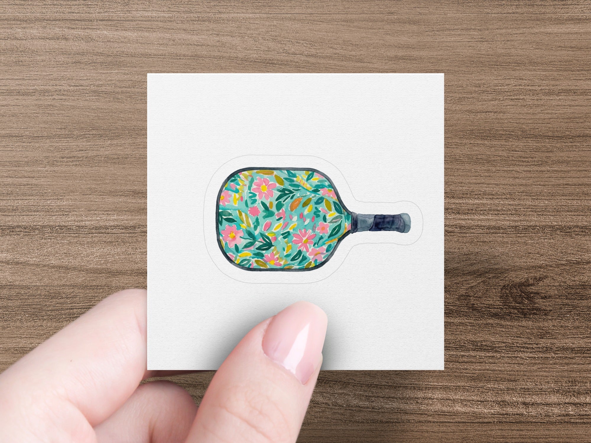 Pickleball paddle with floral pattern held by a hand.