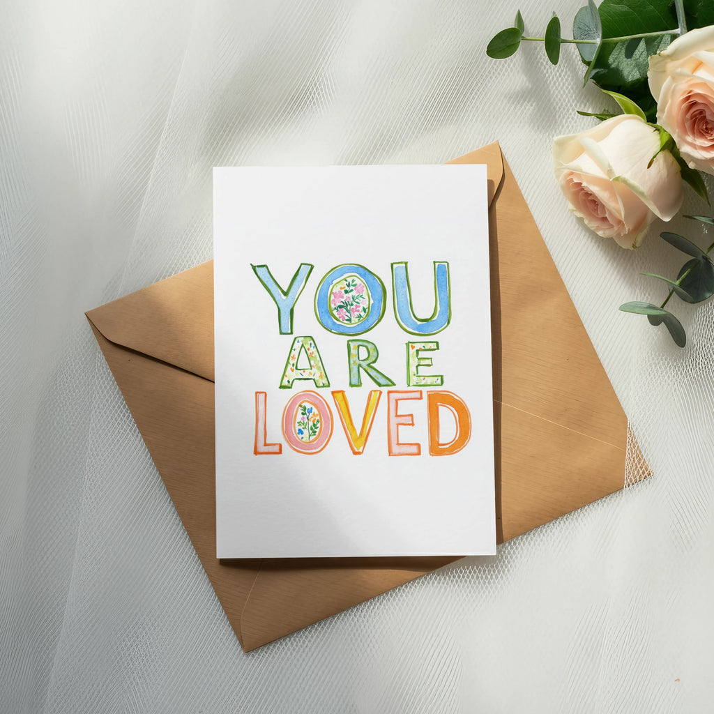 Greeting card with "YOU ARE LOVED" in colorful floral letters on a kraft envelope.
