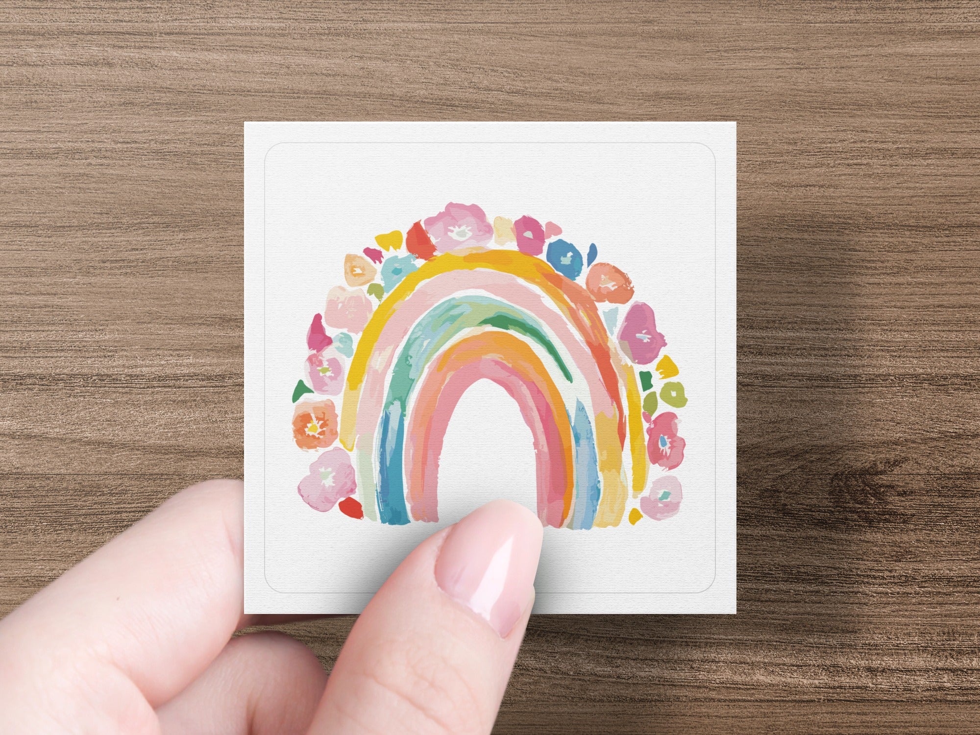 Hand holding a square card with a watercolor rainbow and floral border.