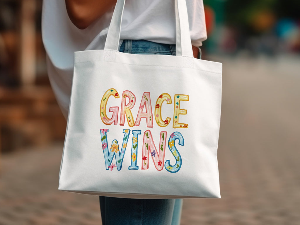 White canvas tote bag with colorful "GRACE WINS" text and floral accents.