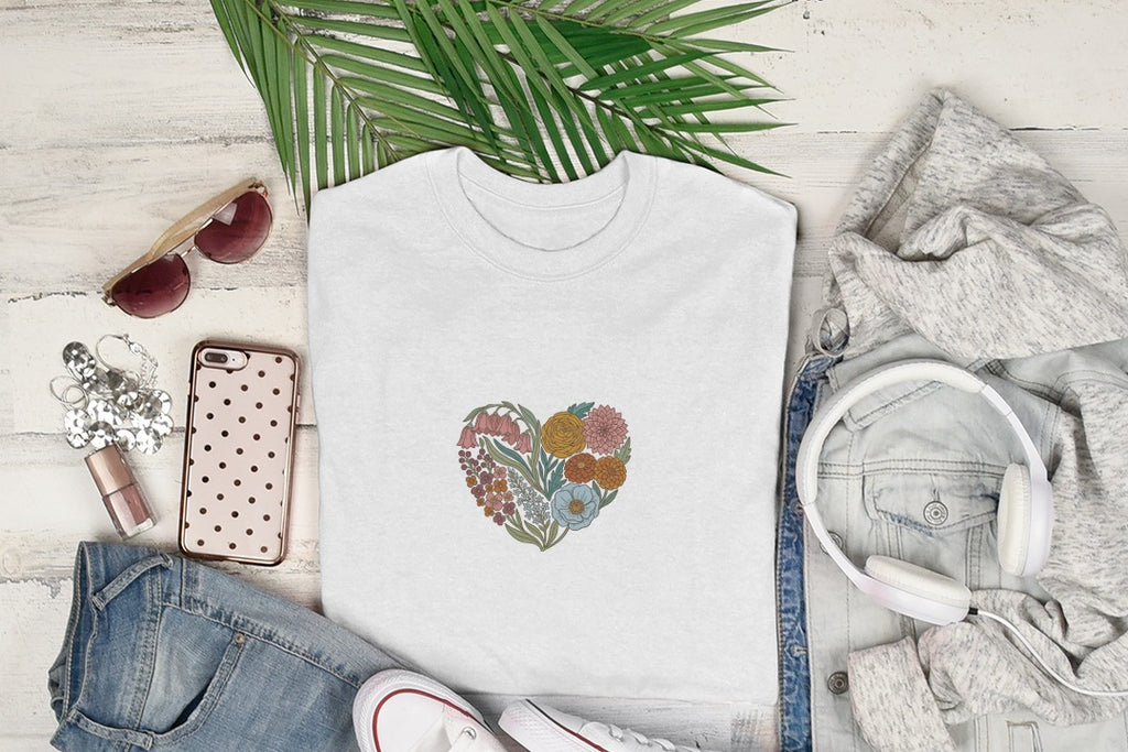 White t-shirt with floral heart design, surrounded by clothing and accessories.