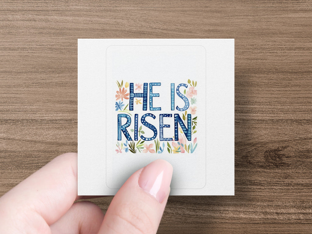 "He is Risen" text with floral accents on a sticker held by a hand.