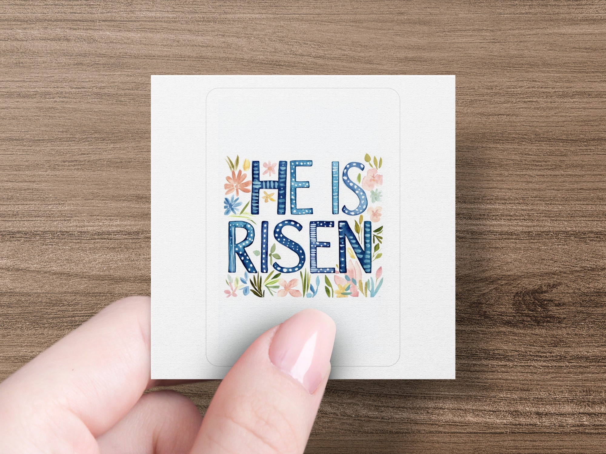"He is Risen" text with floral accents on a sticker held by a hand.