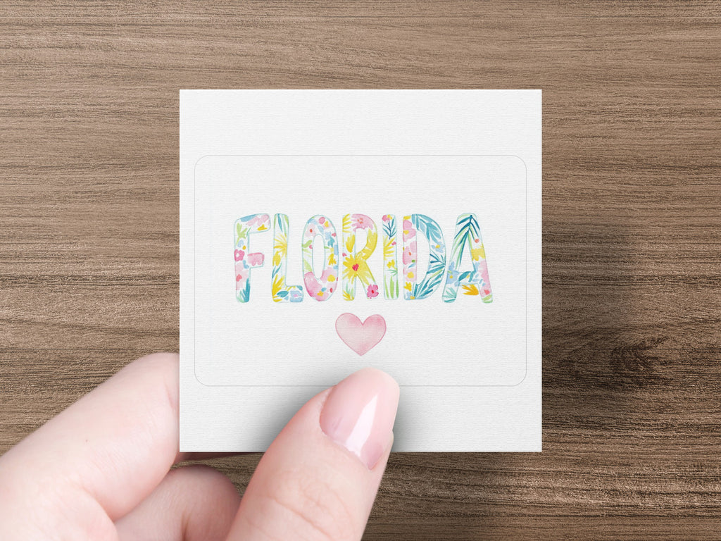 Hand holding square card with "FLORIDA" in colorful watercolor letters and a pink heart.
