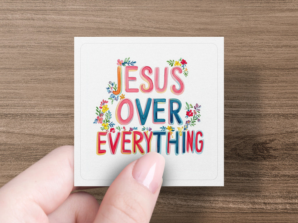 "JESUS OVER EVERYTHING" text with floral accents on a white card held by a hand.