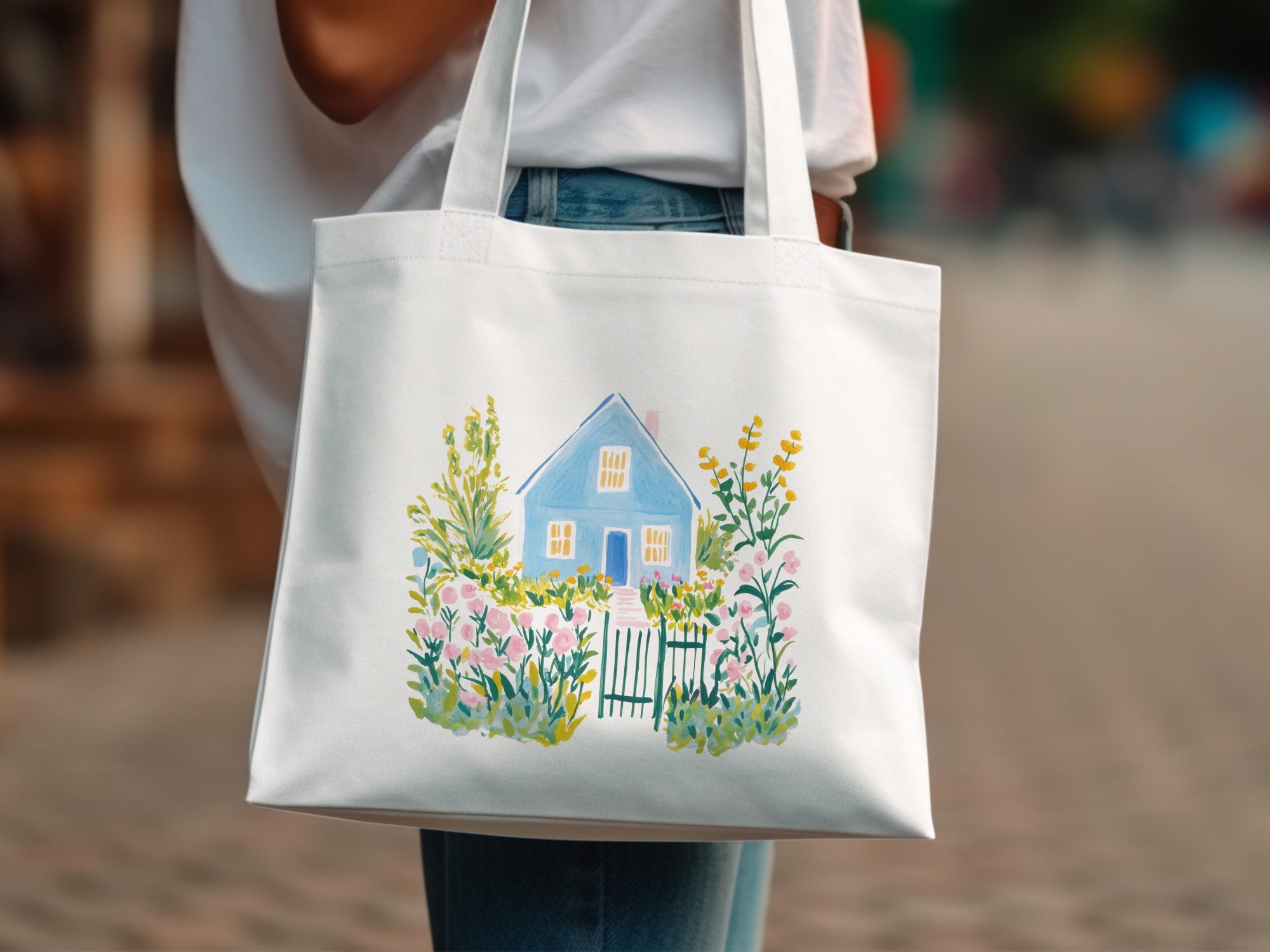 White tote bag with watercolor painting of a blue house with a pink flower garden.