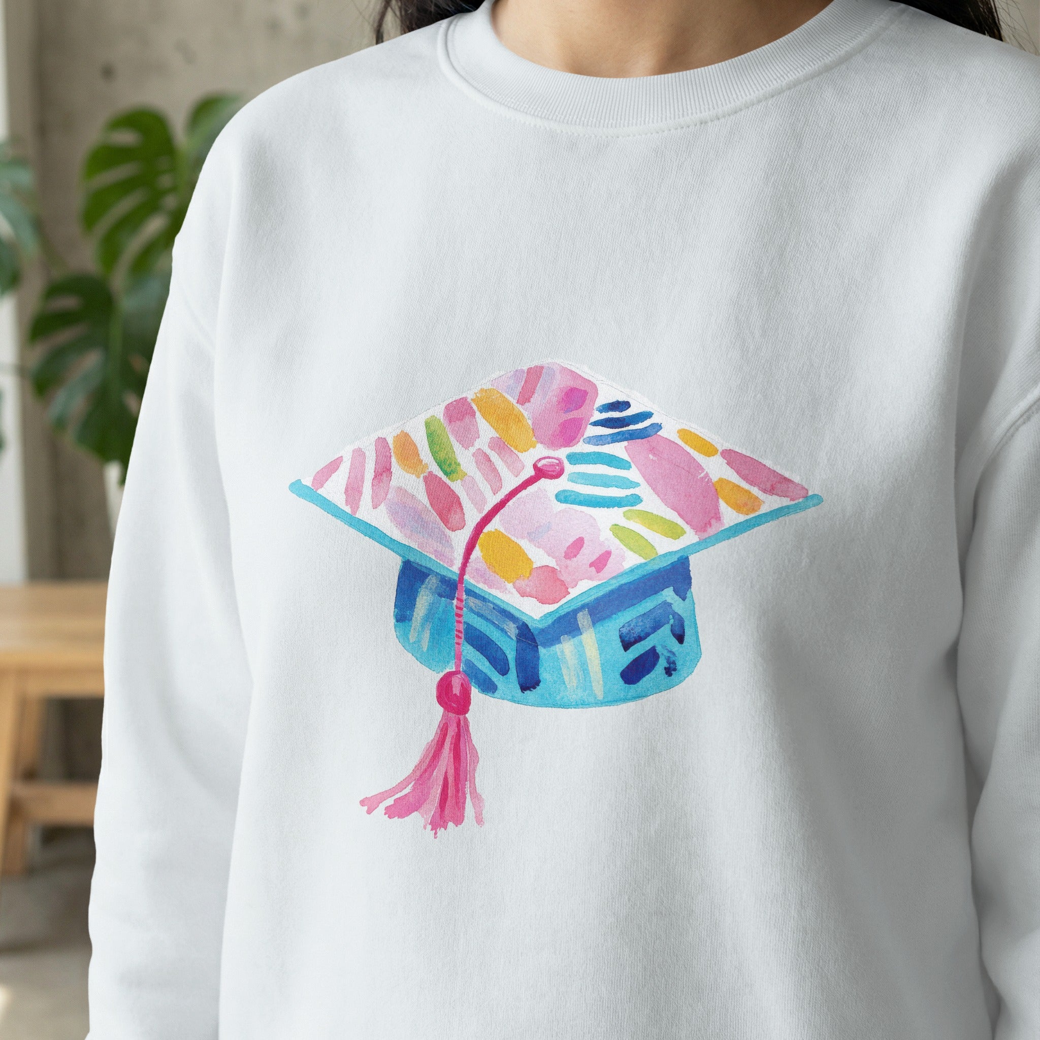 White sweatshirt featuring a watercolor graduation cap with pink and blue accents.