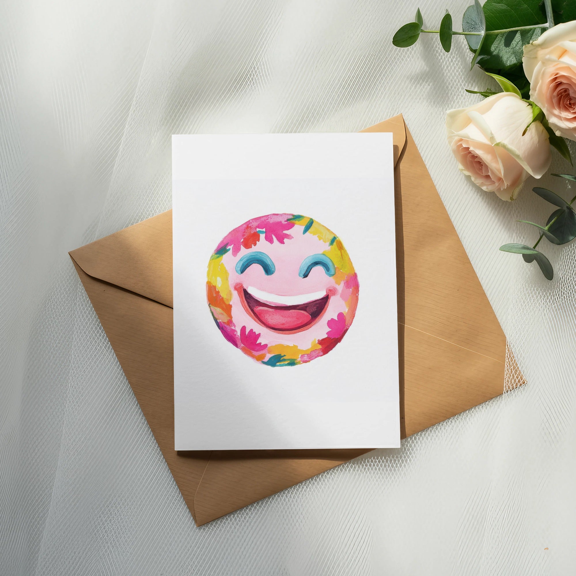 Watercolor smiley face with flowers on a card atop a brown envelope and roses.