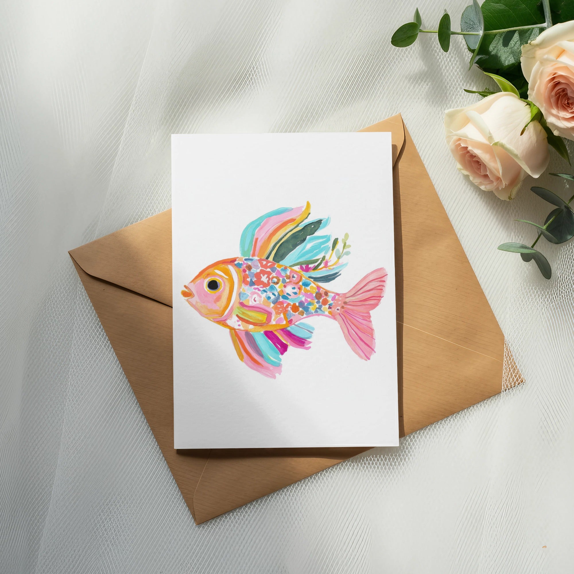 Colorful watercolor fish illustration on white card resting on brown envelope with roses.