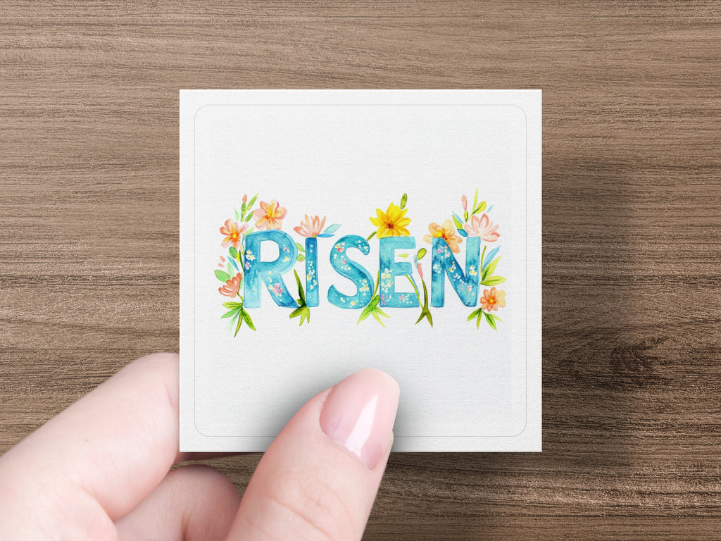 "Risen" text adorned with watercolor flowers held by a hand.