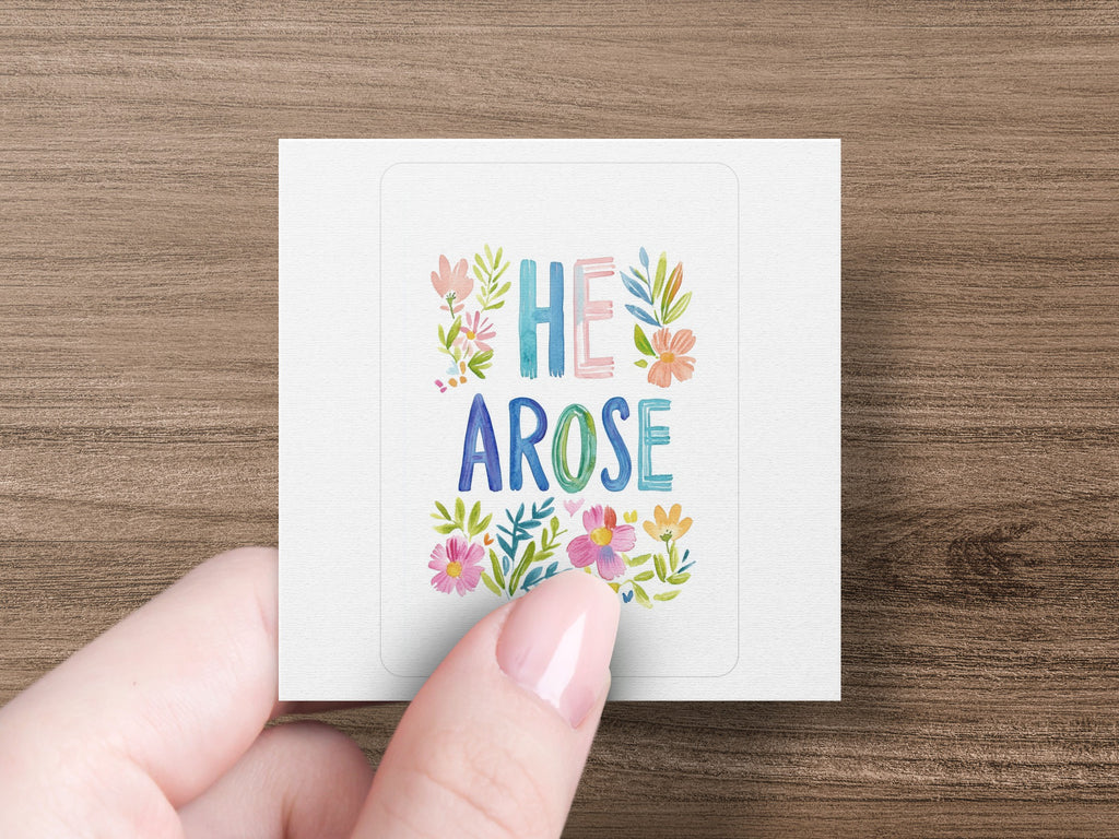 Square sticker with "HE AROSE" in colorful floral lettering held by a hand.