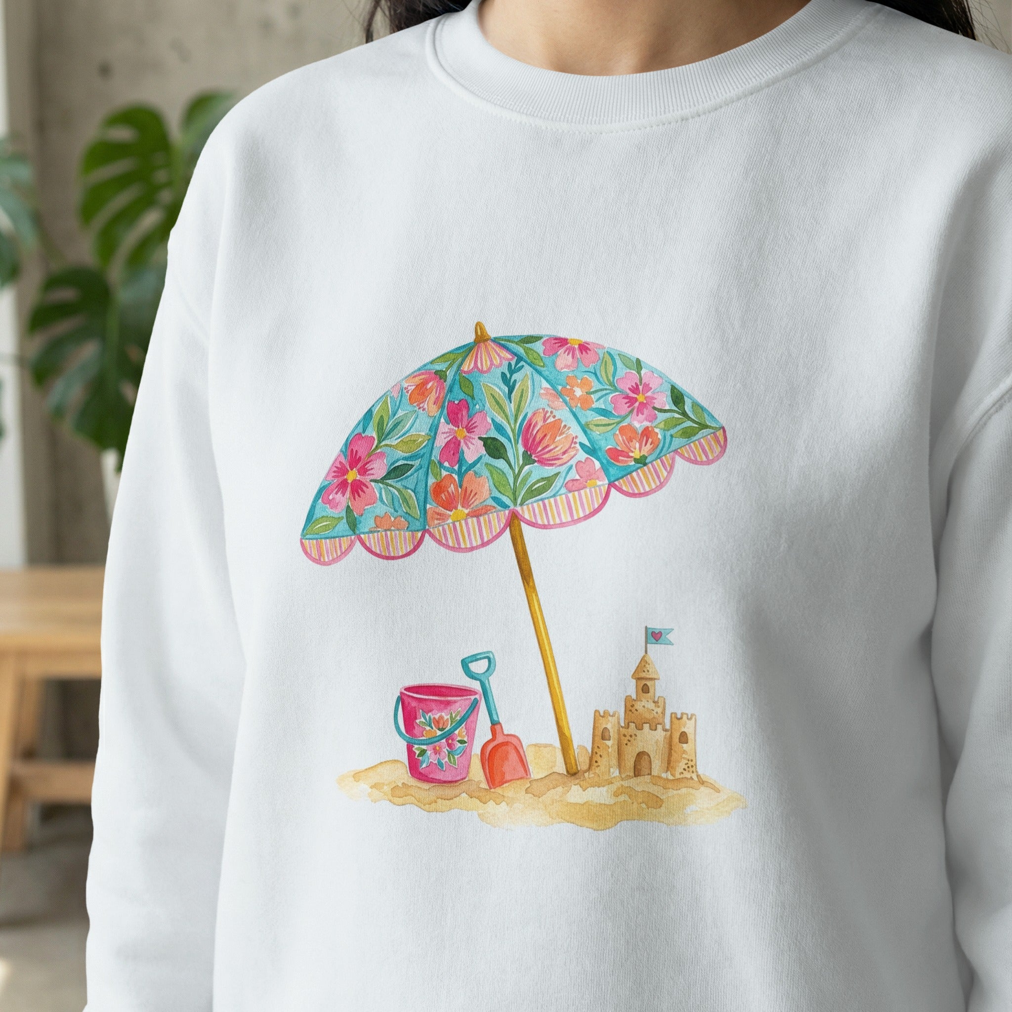 White sweatshirt with a watercolor beach scene: umbrella, sandcastle, and toys.
