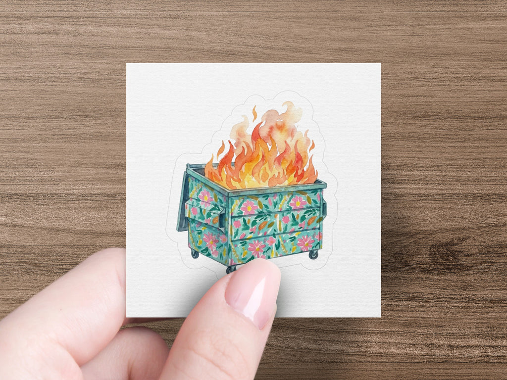 Hand holding a sticker of a teal dumpster with floral pattern on fire.
