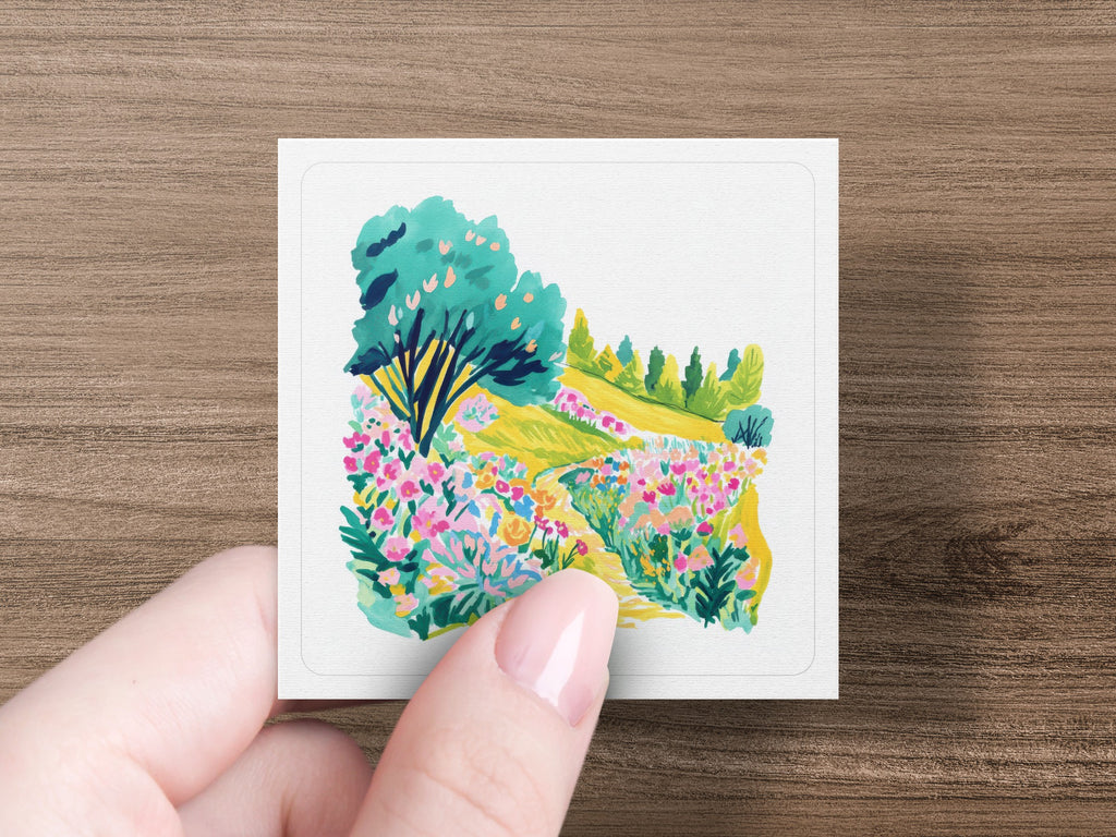 Hand holding a square card with a vibrant painting of a garden path.
