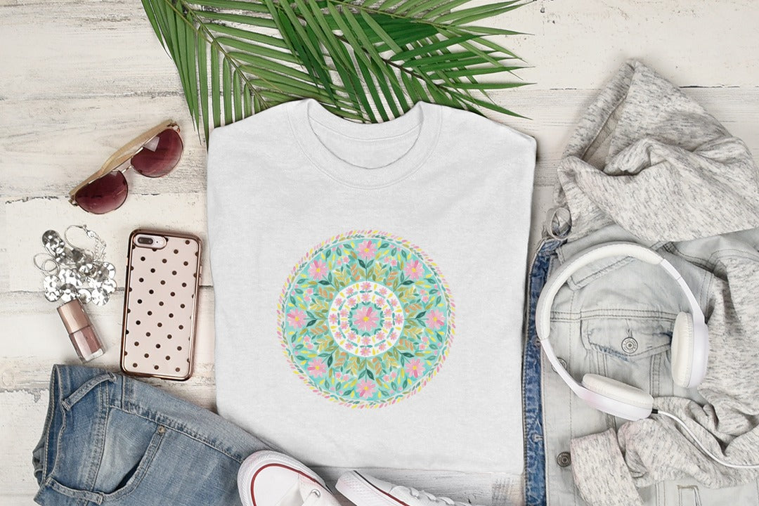White t-shirt with colorful mandala design, next to jeans, sunglasses, and headphones.
