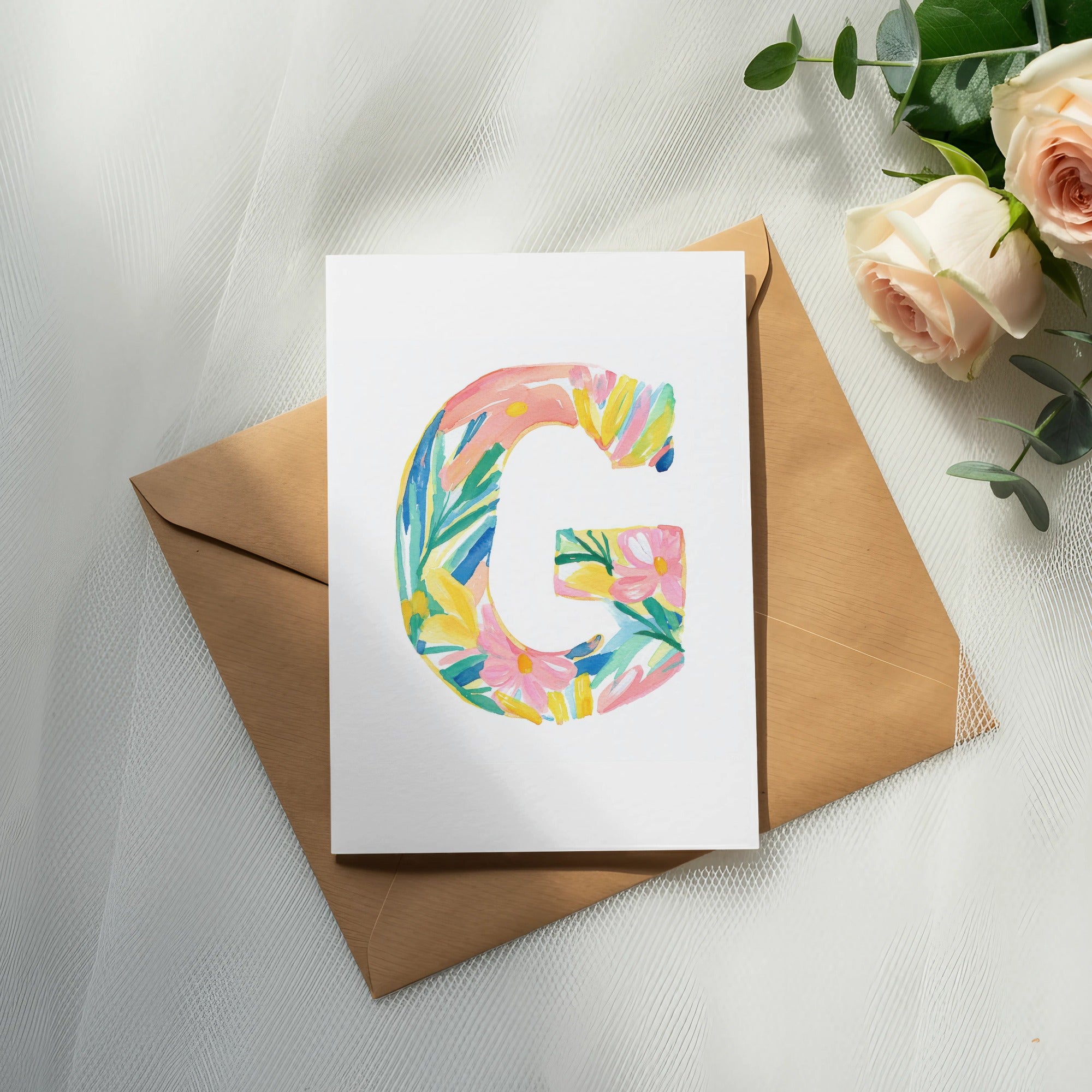 Watercolor letter G with flowers and leaves on cardstock, next to roses.