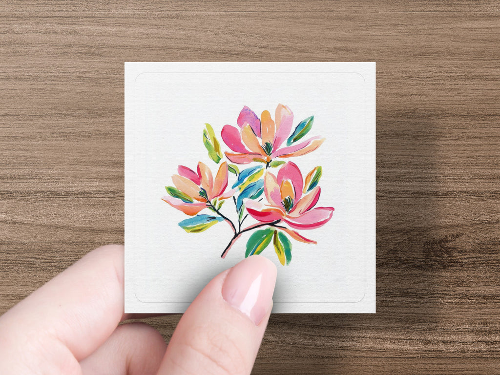 Three watercolor magnolias with green leaves on a white card held by a hand.