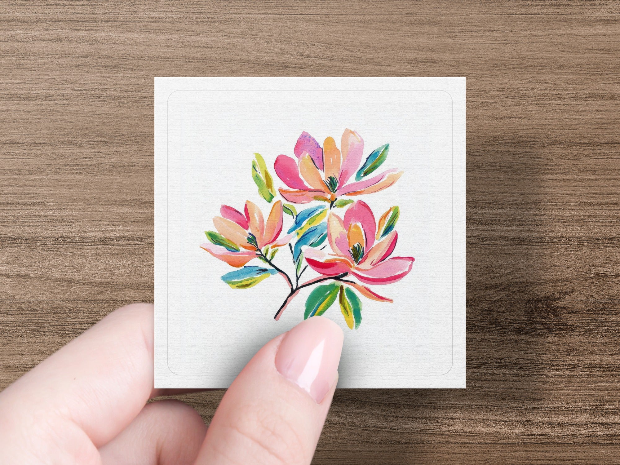 Three watercolor magnolias with green leaves on a white card held by a hand.