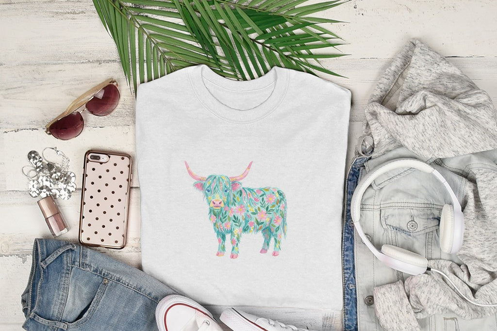 White t-shirt with a floral highland cow graphic, displayed with sunglasses, jeans, and headphones.