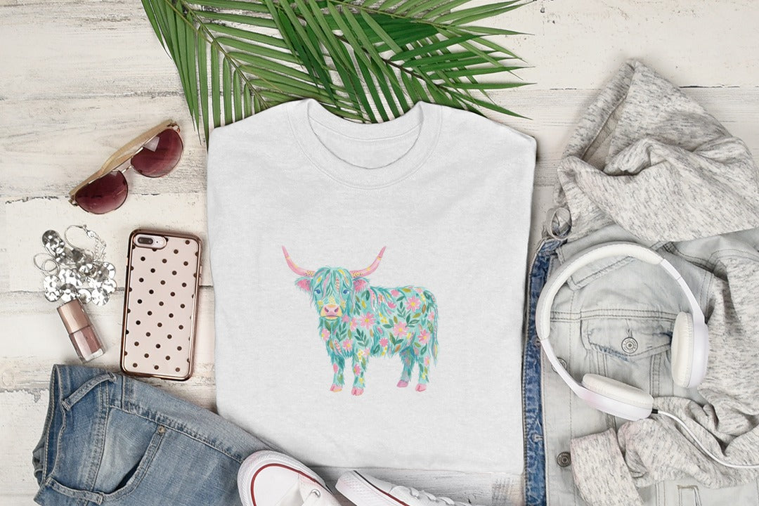 White t-shirt with a floral highland cow graphic, displayed with sunglasses, jeans, and headphones.