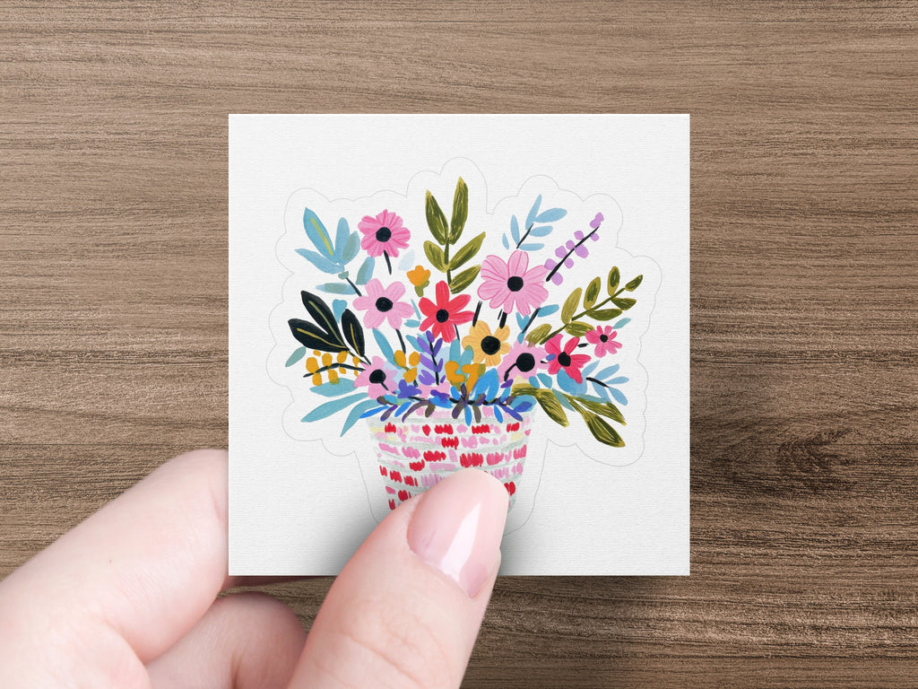 Hand holding a square sticker with a colorful floral arrangement in a striped basket.