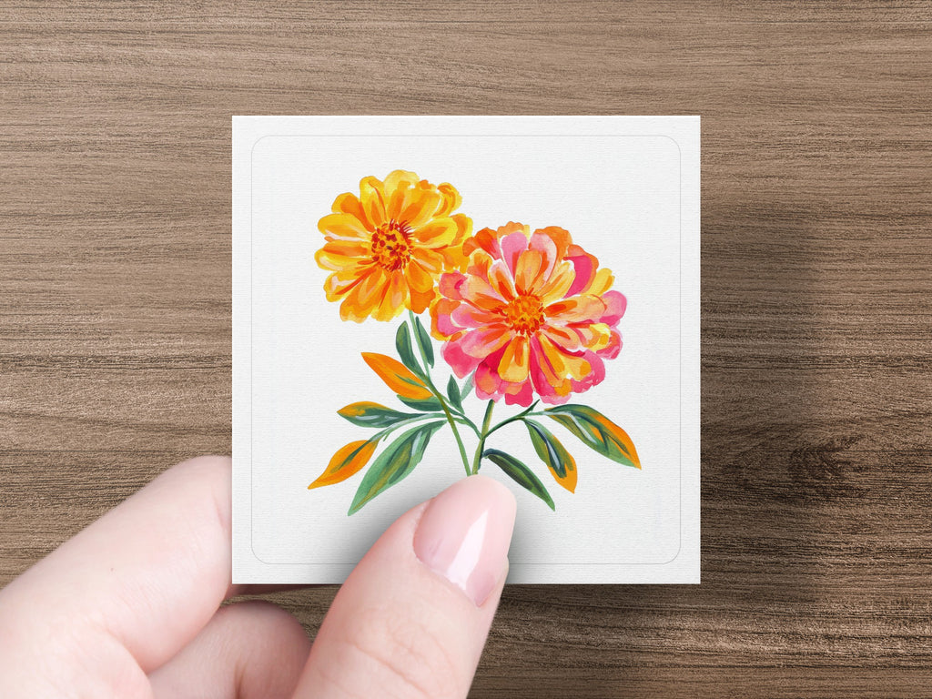 Two vibrant watercolor marigolds in orange and pink on a white card held by a hand.