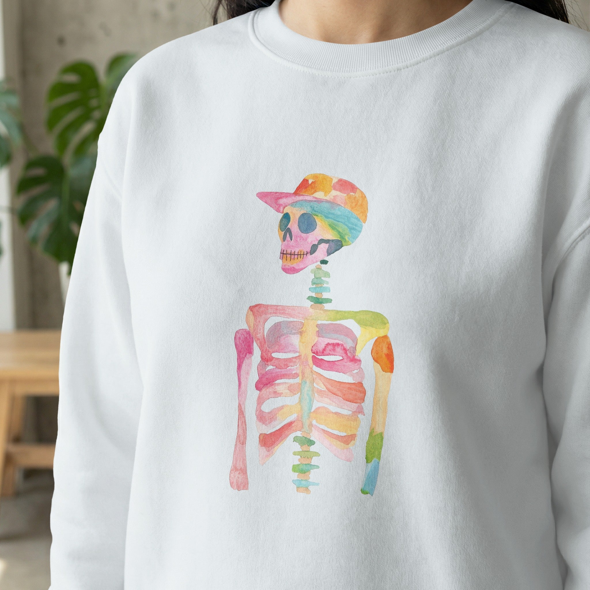 Watercolor skeleton wearing a colorful baseball cap on a white sweatshirt.