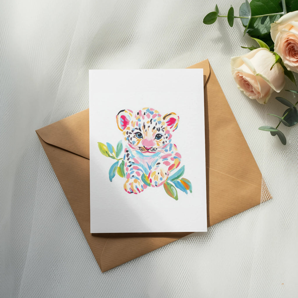 Watercolor painting of a colorful baby leopard cub on a card atop a brown envelope.