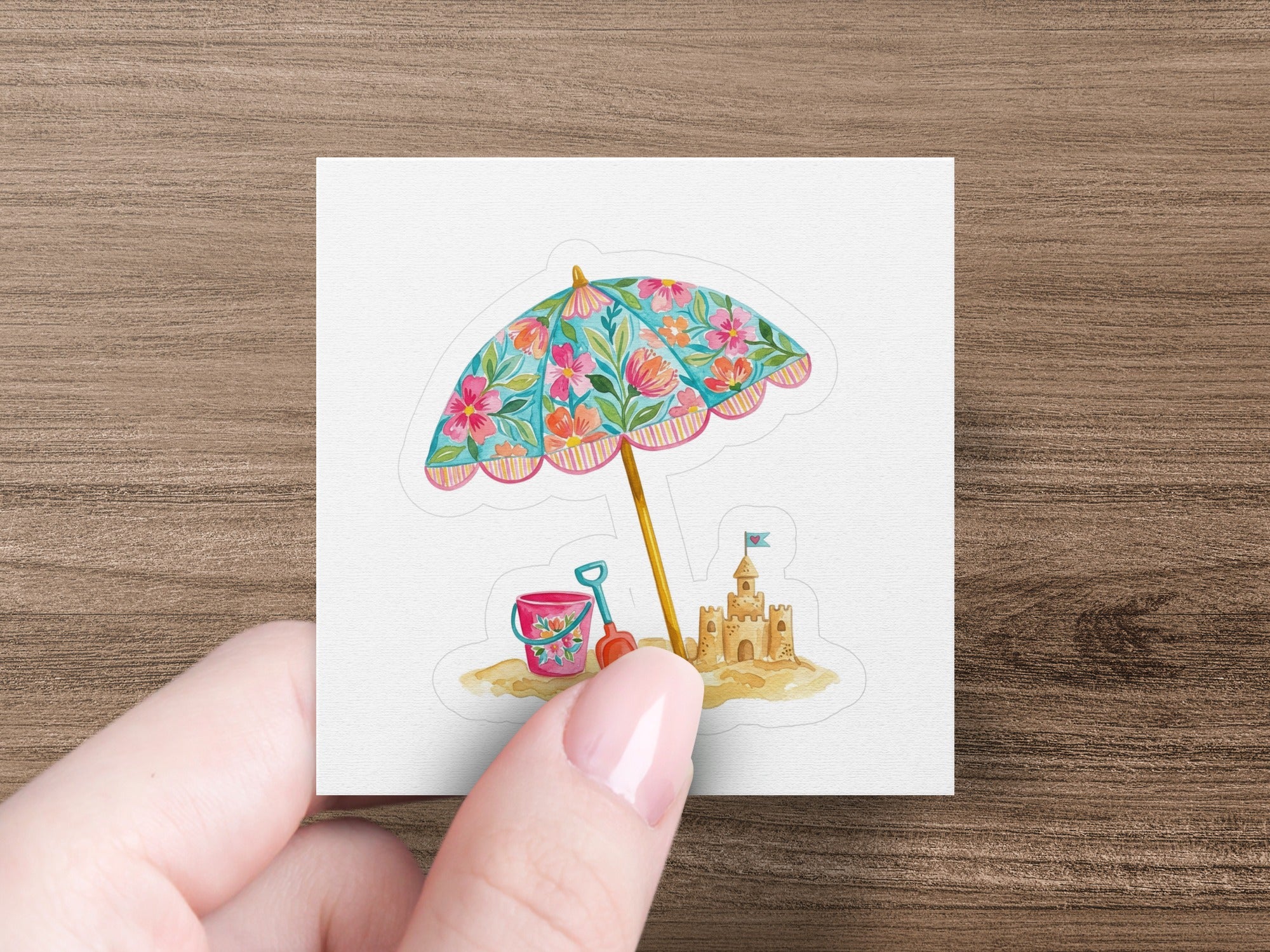 Hand holding a sticker of a beach umbrella, sandcastle, shovel, and pail.