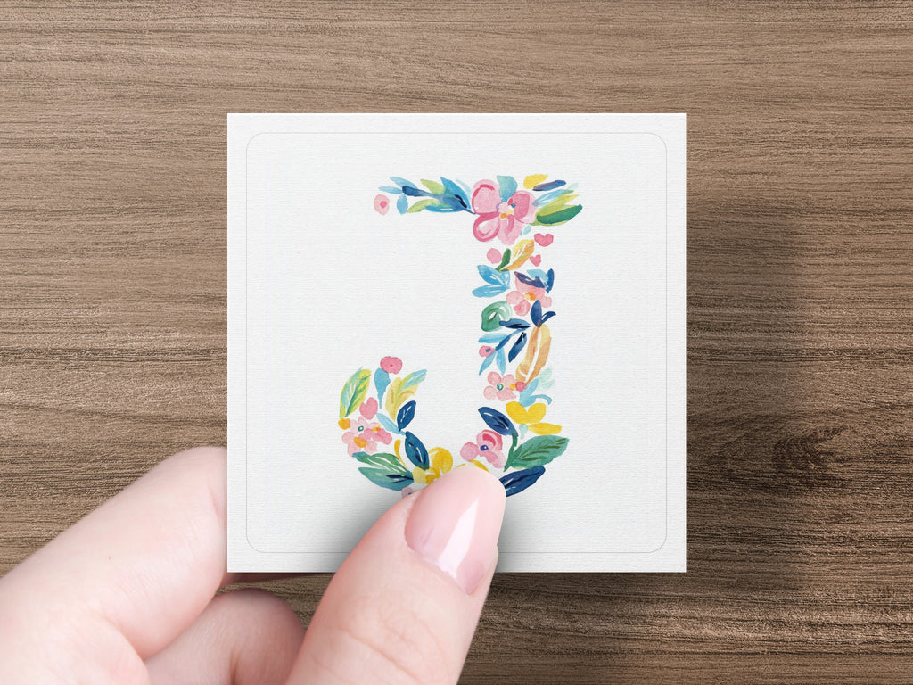 Floral letter J on a white card held by a person's fingers.