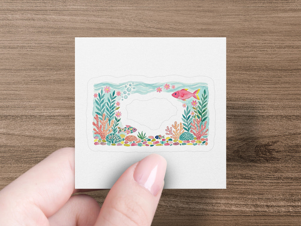 Hand holding a square sticker featuring a colorful coral reef with fish.