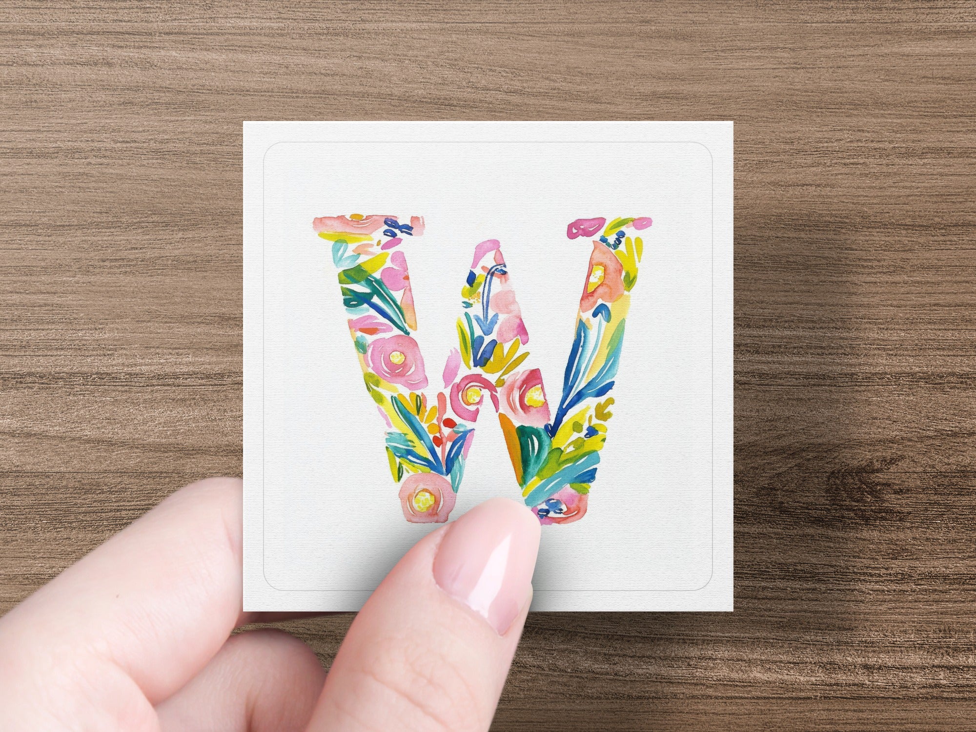 Hand holding a square card with a floral letter W.