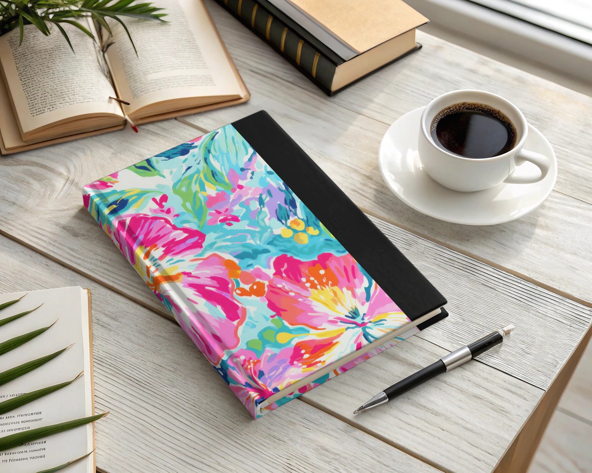 Colorful floral notebook with black spine and pen, next to coffee cup.