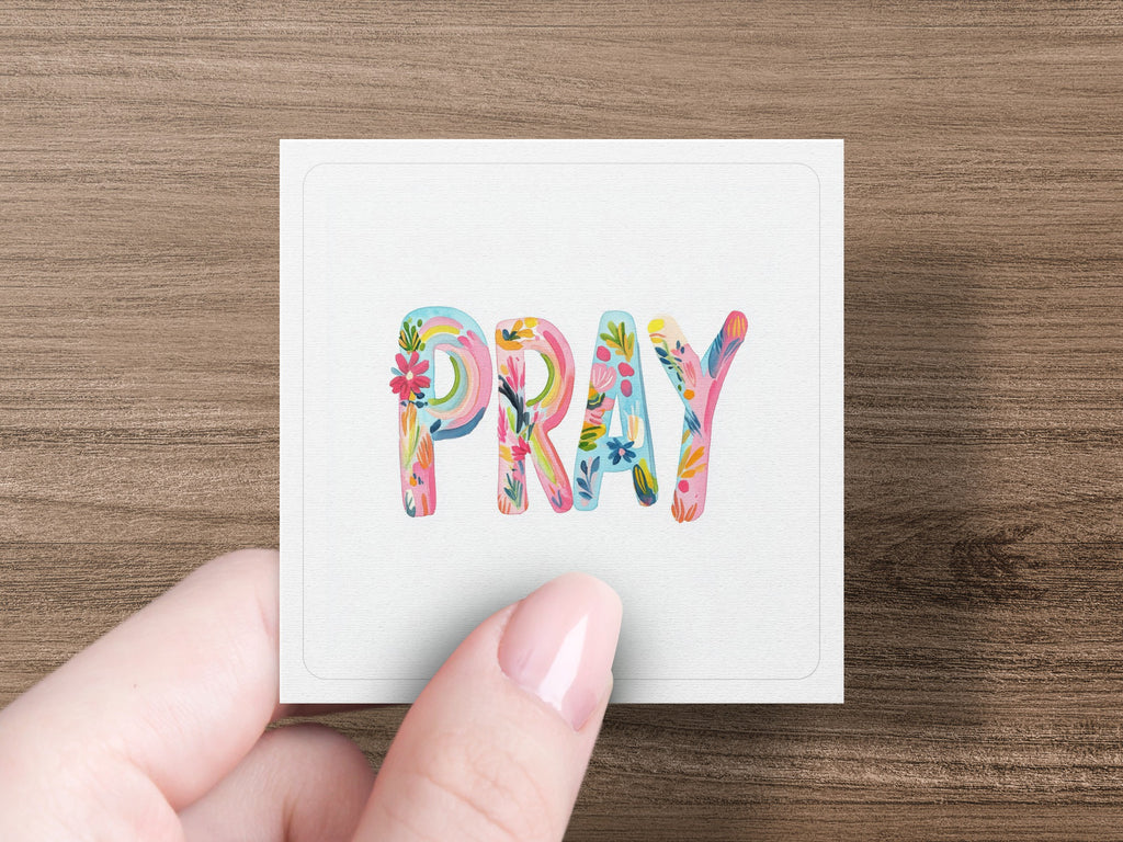 White card with "PRAY" in colorful floral letters held by a hand.