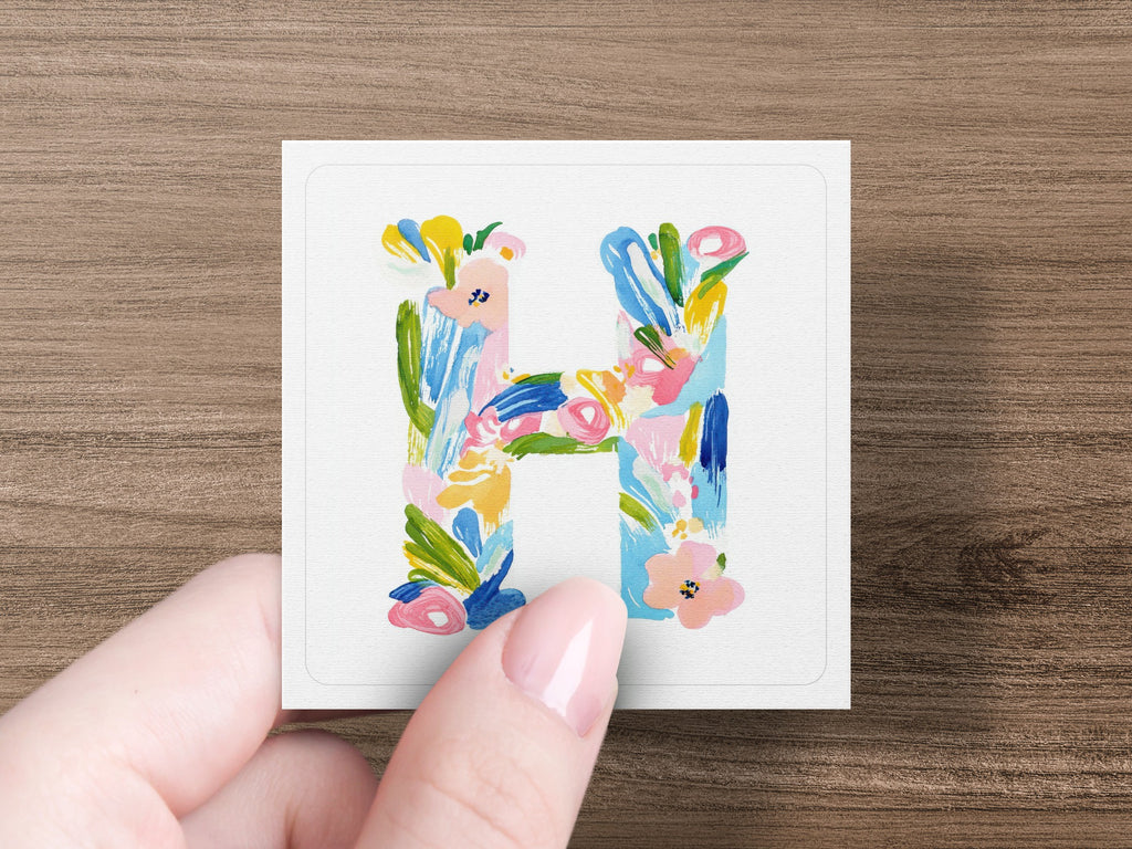 Hand holding a square card with a watercolor letter "H" design.