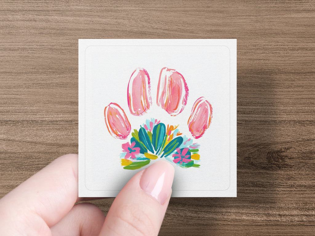Pink paw print above colorful flowers on a white card held by a hand.