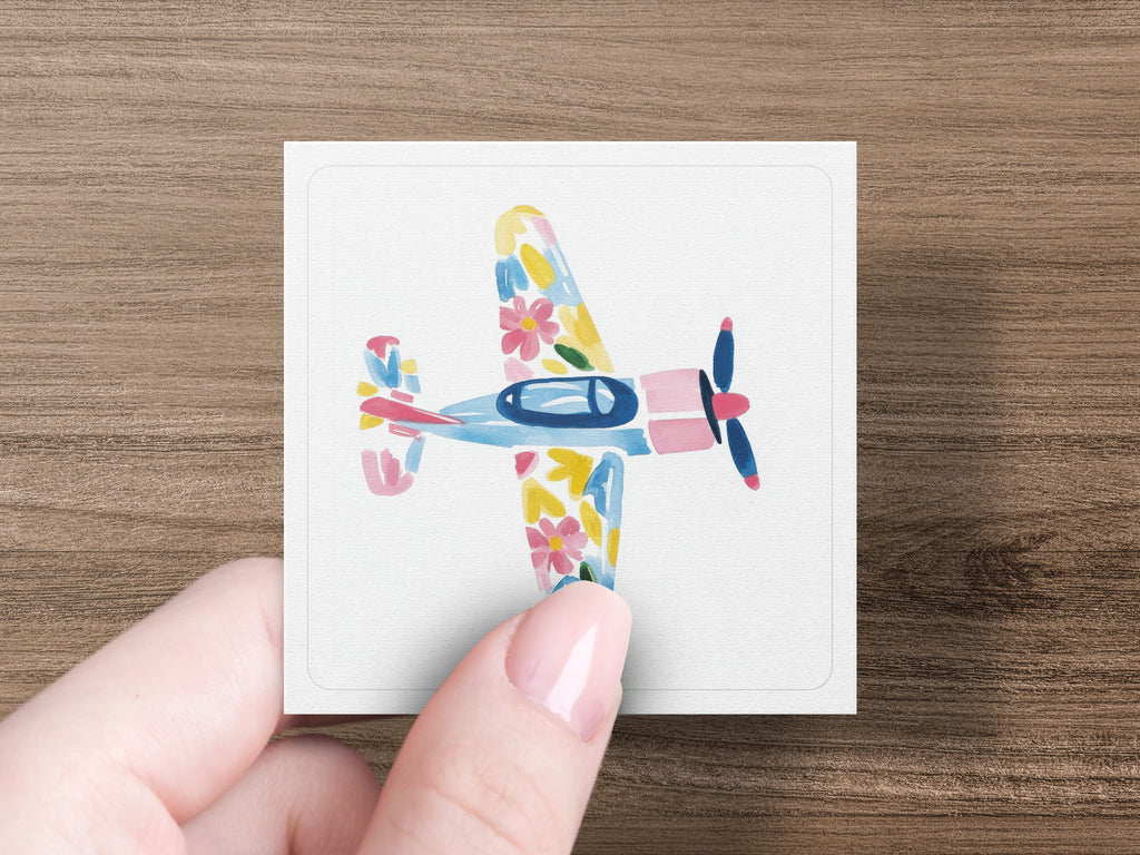 Hand holding a square card featuring a watercolor airplane with floral patterns.