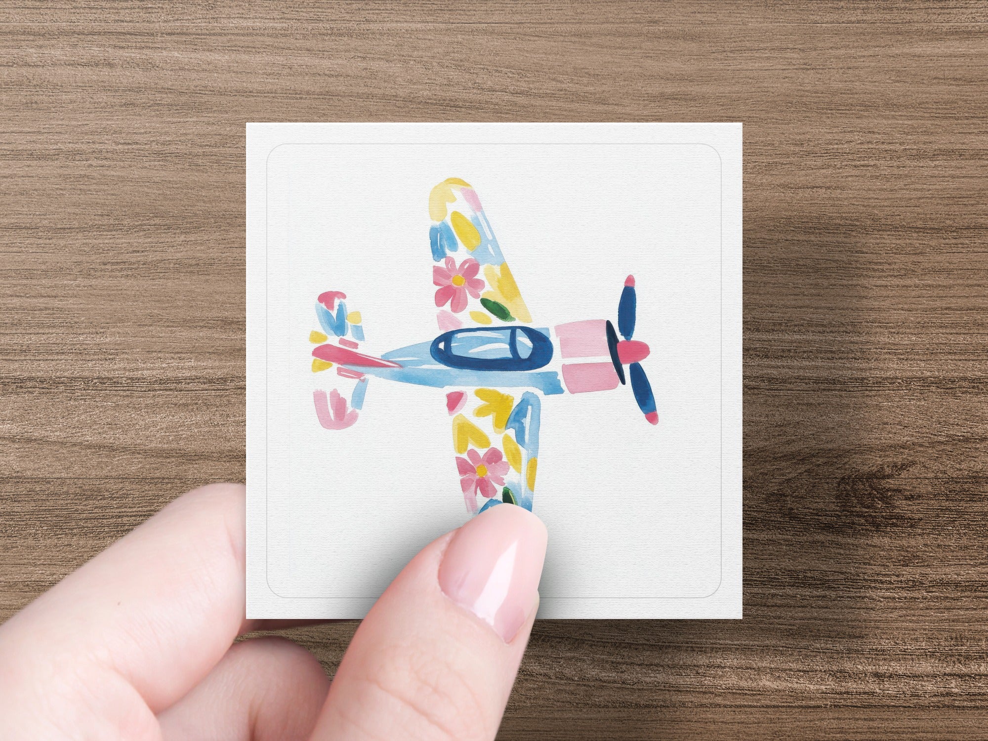 Hand holding a square card featuring a watercolor airplane with floral patterns.