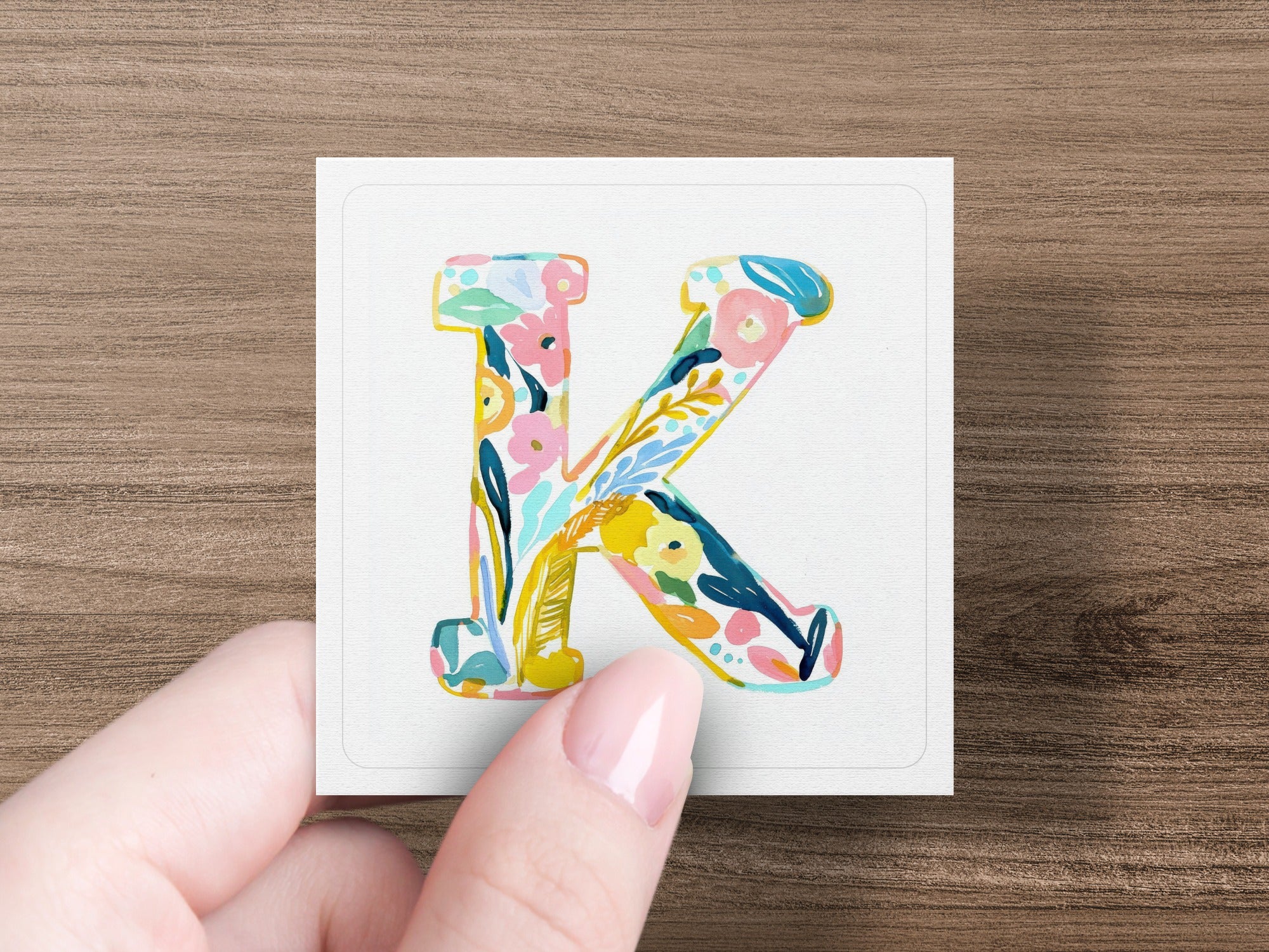 A hand holds a square tile with a colorful floral letter K.