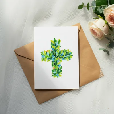 Watercolor cross with green leaves and blue accents on card, with envelope and roses.