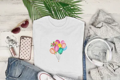 White t-shirt with colorful watercolor balloons graphic, laid flat with sunglasses, phone, jeans, and headphones.