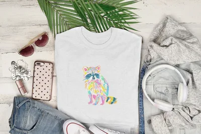 White t-shirt with colorful watercolor raccoon graphic, surrounded by clothing and accessories.