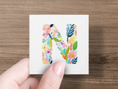 Floral letter N in watercolor on square card held by fingers.