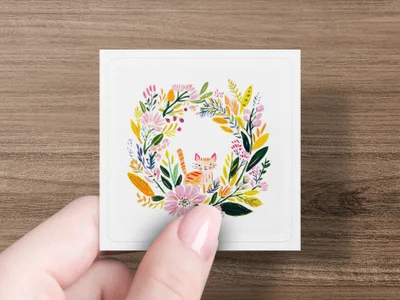 Square card with floral wreath and smiling cat illustration held by a hand.