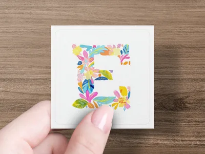 Floral letter E on a white square card held by a hand.