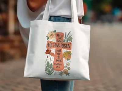 White tote bag with floral cross and "He is not here; HE HAS RISEN" text.