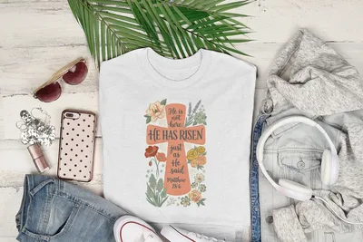 White t-shirt with "He Has Risen" and floral cross graphic, Matthew 28:6.