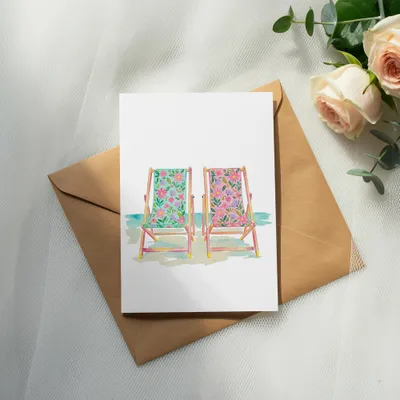 Watercolor painting of two floral deck chairs by a blue ocean.
